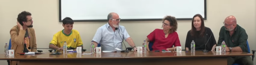 A imagem mostra uma mesa-redonda com seis pessoas sentadas atrás de uma mesa comprida de madeira. Todas têm microfones à frente e garrafas de água. Ao fundo, há uma parede clara com uma tela branca de projeção. À esquerda, um homem de óculos, barba curta e paletó marrom olha para o lado. Ao lado dele, uma pessoa veste uma camisa amarela da seleção brasileira e segura papéis. No centro, um homem grisalho de óculos e camisa clara conversa com uma mulher de óculos e blusa vermelha. À direita, uma mulher de cabelos castanhos longos e blusa preta sorri, enquanto um homem de barba grisalha e camisa verde olha para ela. Trata-se de um momento de discussão entre os participantes.